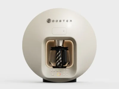Cream DOSTEA beverage machine with rounded circular housing, illuminated brewing chamber, glass carafe, and integrated touch control