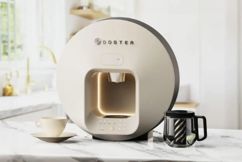 Cream DOSTEA tea machine with illuminated brewing chamber and glass carafe displayed on a marble kitchen counter.