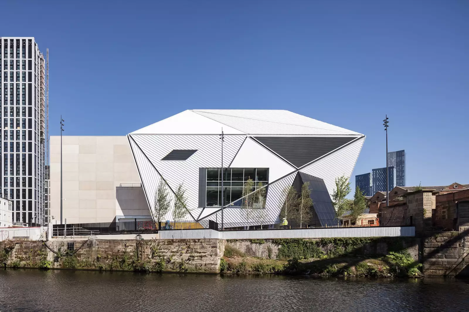 Angular white contemporary building with geometric metal cladding beside a canal, framed by modern high-rise towers under a clear blue sky.