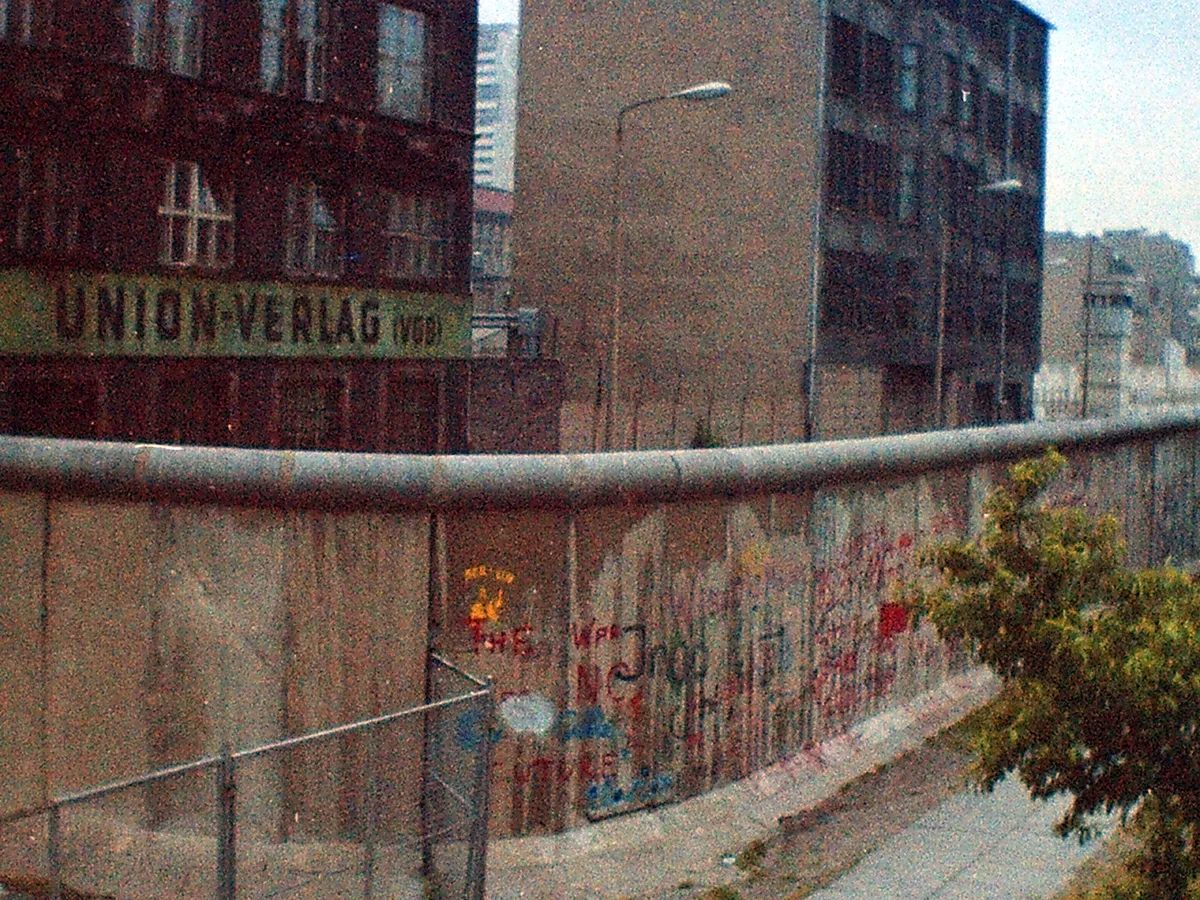Grainy urban streetscape with graffiti-covered Berlin Wall, industrial buildings, and a faded UNIVERSUM sign overlooking a quiet city street.