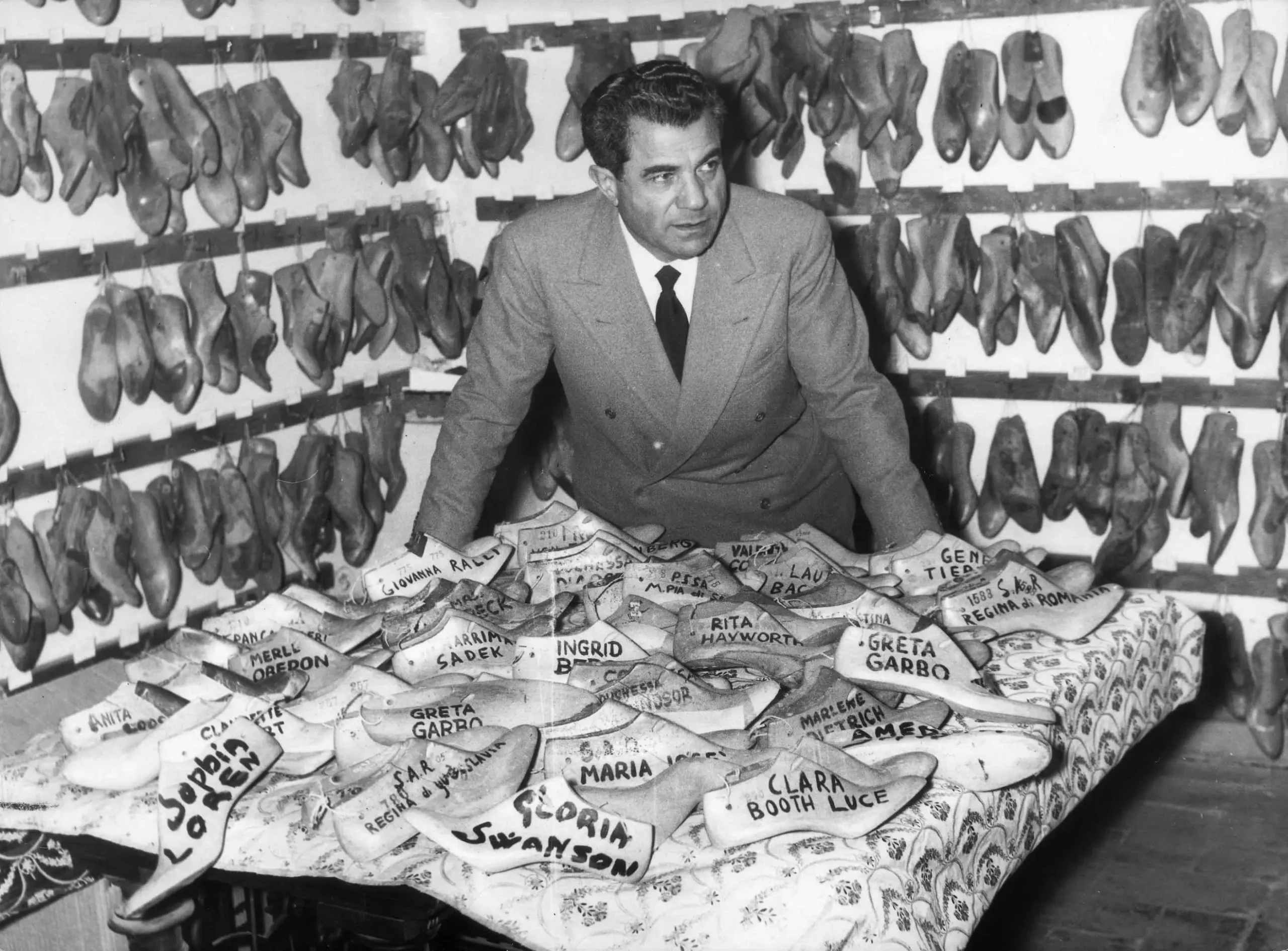 Historic black-and-white photograph of Salvatore Ferragamo standing behind a worktable covered with wooden shoe lasts labeled for notable clients, surrounded by shelves filled with handcrafted lasts inside his workshop.