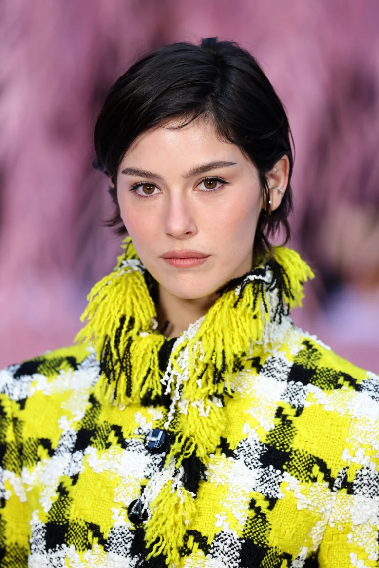 Close-Up Portrait Of Gracie Abrams With Short Dark Hair Wearing A Textured Yellow, Black, And White Woven Jacket With Fringe Detailing Against A Soft Pink Background