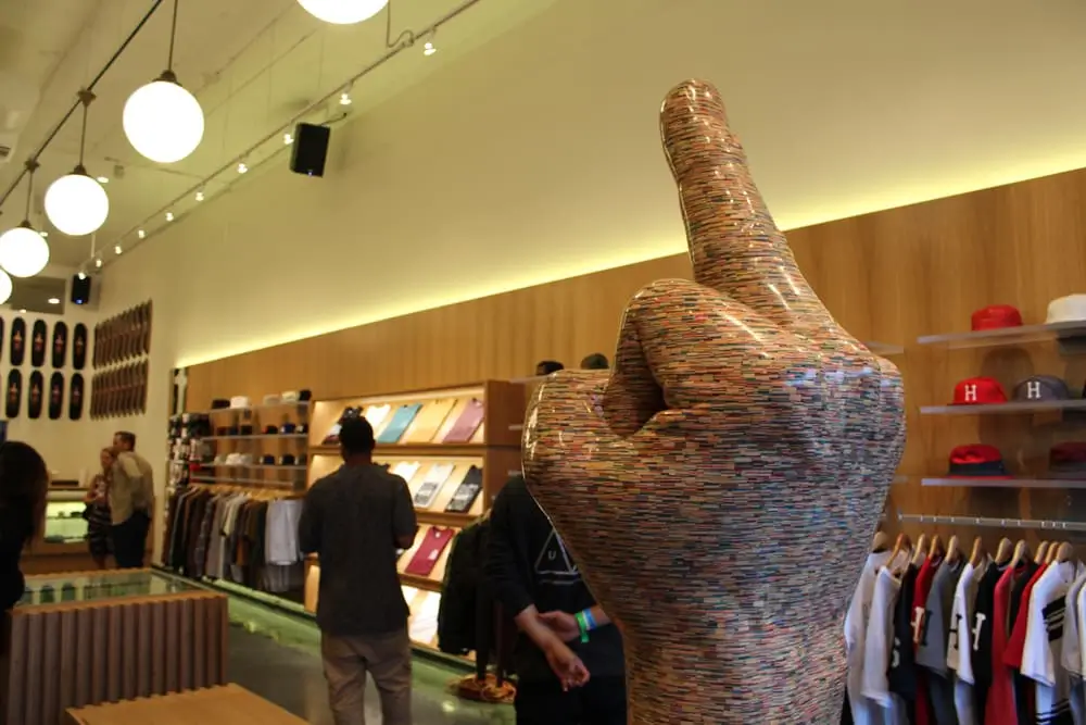 Interior of a HUF retail store featuring a large sculptural hand with a raised middle finger made from stacked skateboard decks, surrounded by apparel displays, hats, and skate-inspired merchandise.