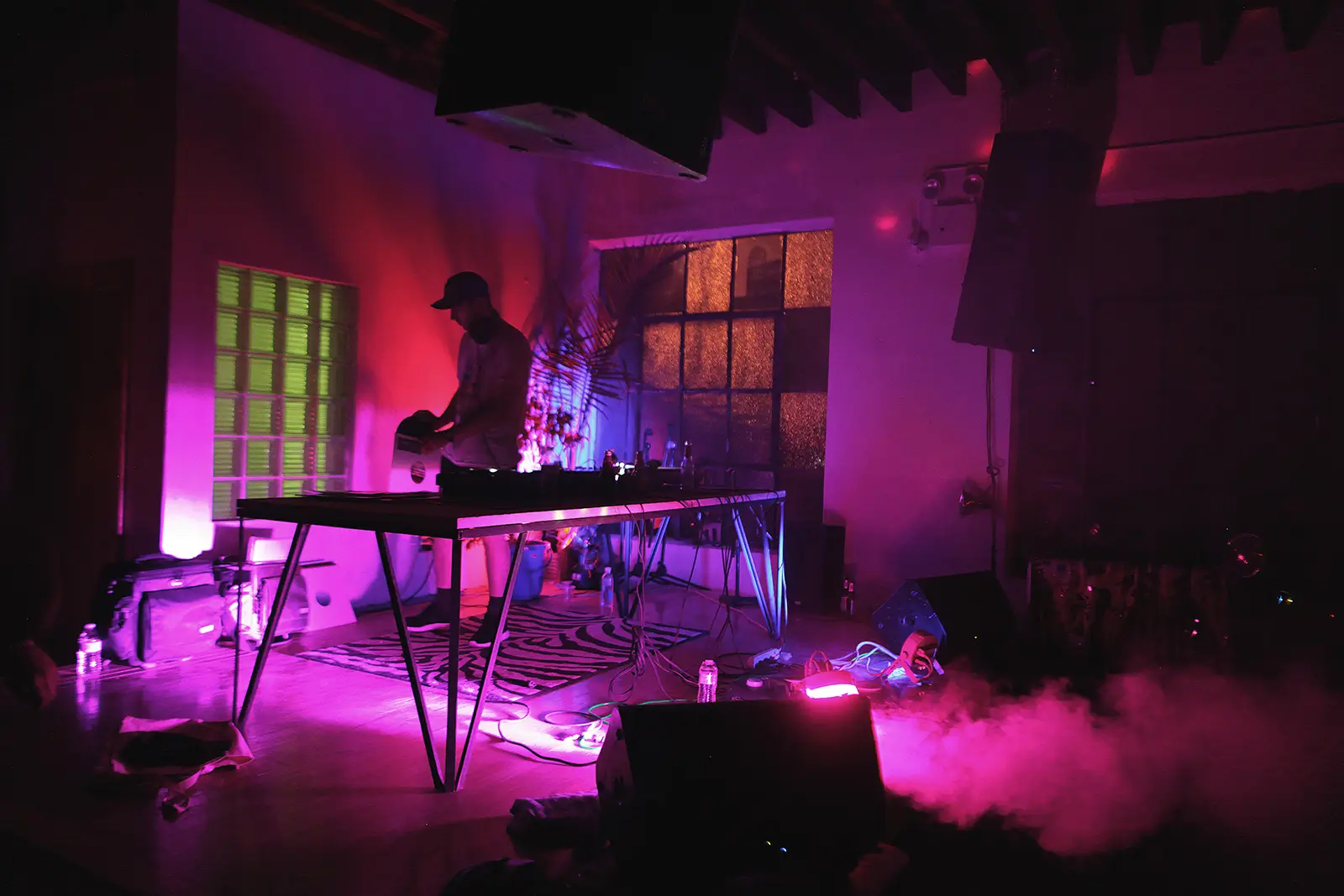 Atmospheric DJ set inside a dimly lit venue with magenta and violet lighting, a minimalist performance table, drifting fog, large speakers, and industrial windows creating an intimate late-night club atmosphere.