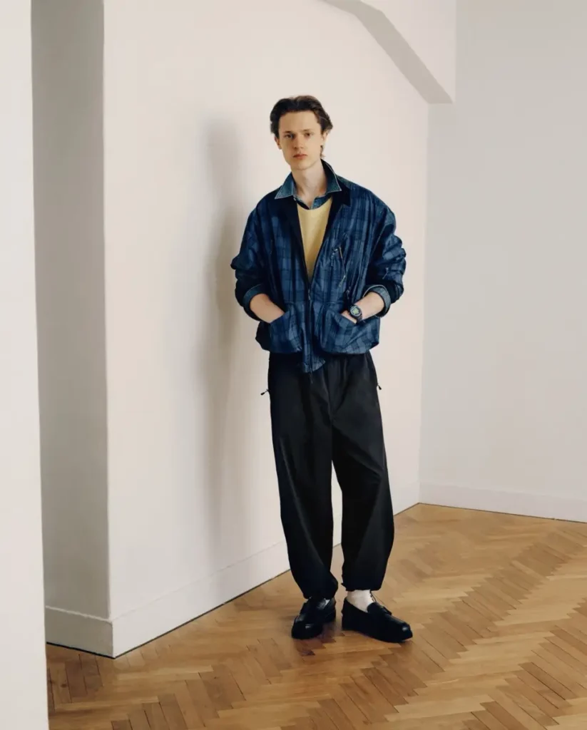 Full-body view of model in a blue plaid jacket, loose black trouser and black shoes stand in a min white interior with parquet floor