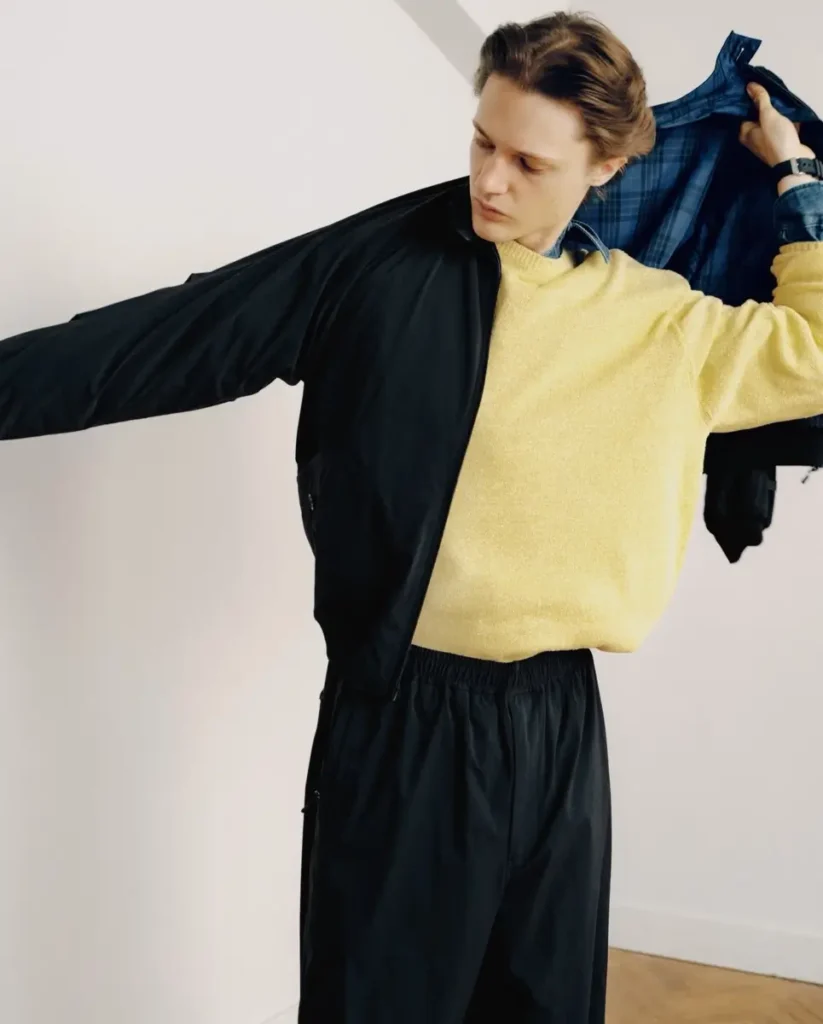 Model remove a reversible blue plaid and black jacket, revealing a pale yellow sweater layered over a denim shirt and loose black trouser