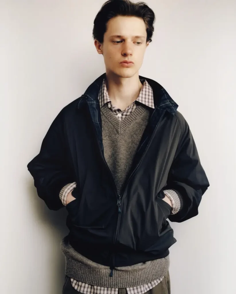 Model wearing a black technical jacket over a gray V-neck knit sweater and check shirt, hands tuck into the jacket pocket