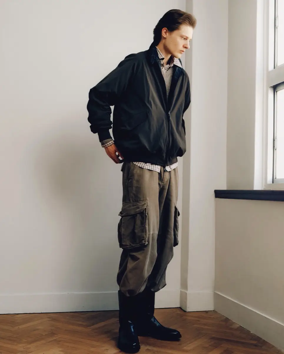 Model wearing a black technical jacket layered over a gray knit sweater and checked shirt with oversized cargo trousers and black shoes beside a window.