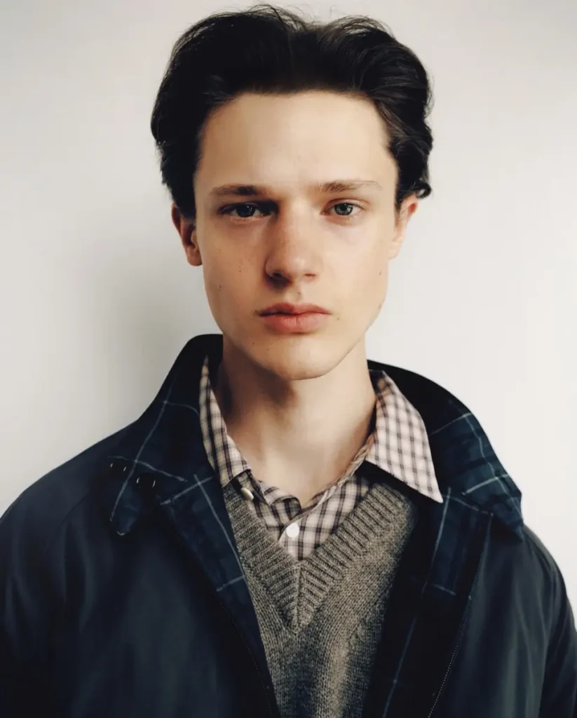 Close-up portrait of model wearing a black jacket with blue plaid lining over a gray knit sweater and check shirt