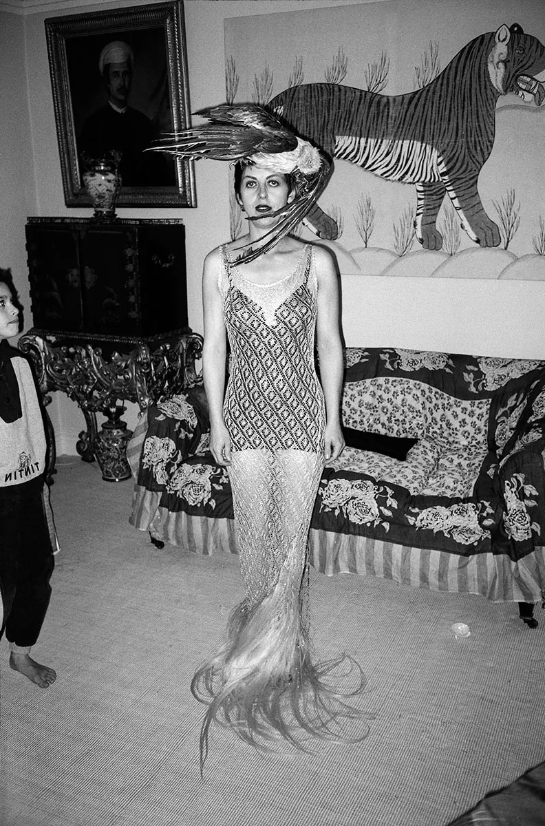 Black-and-white portrait of a woman in a beaded gown and dramatic bird-inspired headpiece standing inside an eclectic, art-filled room.