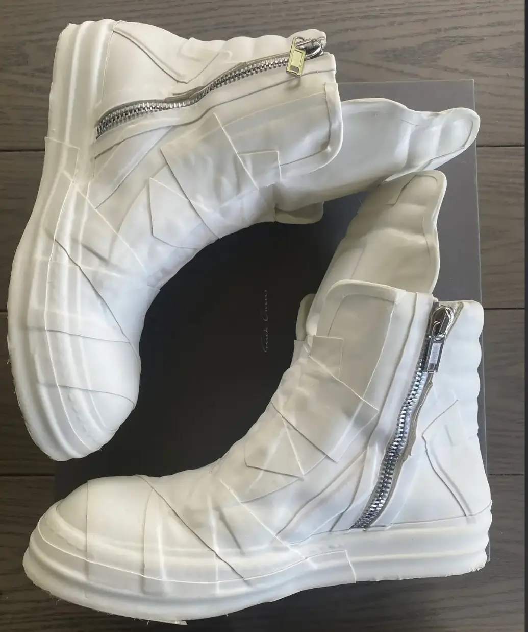 Pair of sculptural white high-top boots with oversized side zippers, layered panel construction, and a chunky wraparound sole, photographed from above on a black shoebox.