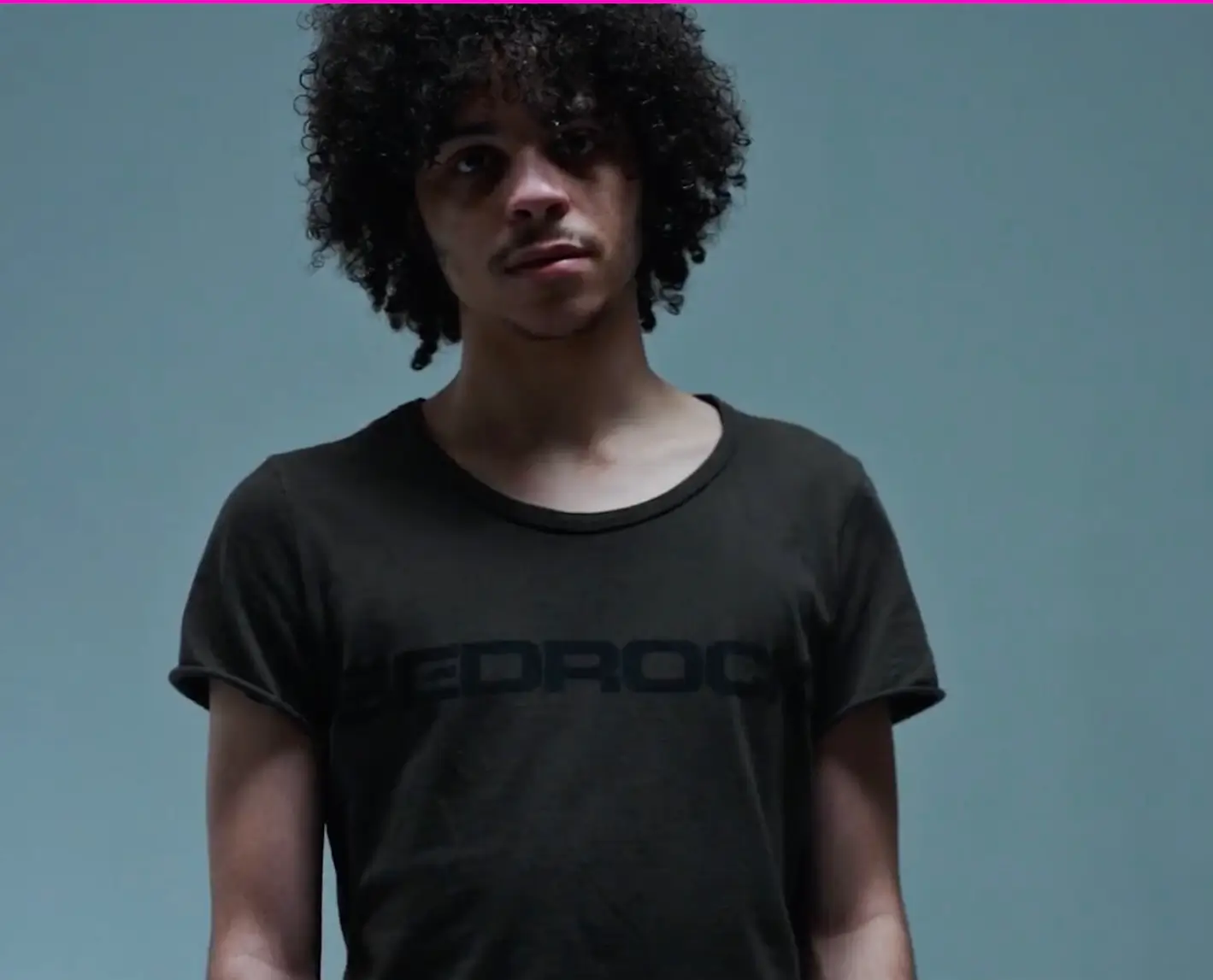 Minimalist portrait of a xaviersobase person with voluminous curly hair wearing a faded black graphic T-shirt, standing against a plain light blue background under soft, subdued studio lighting.