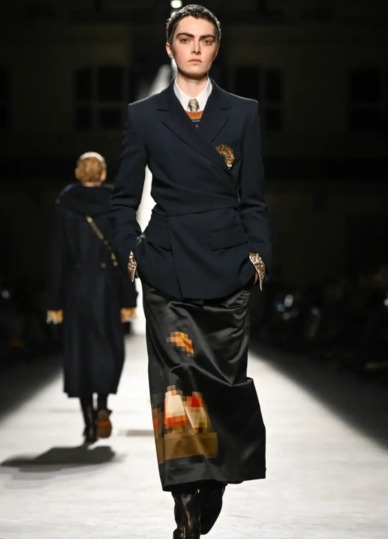 Dries Van Noten runway look featuring a structured navy wrap blazer over a white shirt and tie, paired with a black satin midi skirt decorated with pixelated orange and brown motifs and tall leather boots.