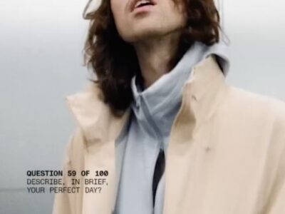 A.G. Cook with shoulder-length brown hair and glasses wearing a beige jacket over a gray hoodie, looking upward against a pale background, with on-screen text reading, “Question 59 of 100: Describe, in brief, your perfect day?”