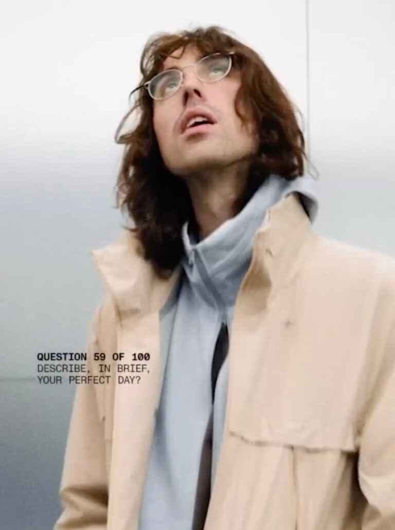 A.G. Cook with shoulder-length brown hair and glasses wearing a beige jacket over a gray hoodie, looking upward against a pale background, with on-screen text reading, “Question 59 of 100: Describe, in brief, your perfect day?”