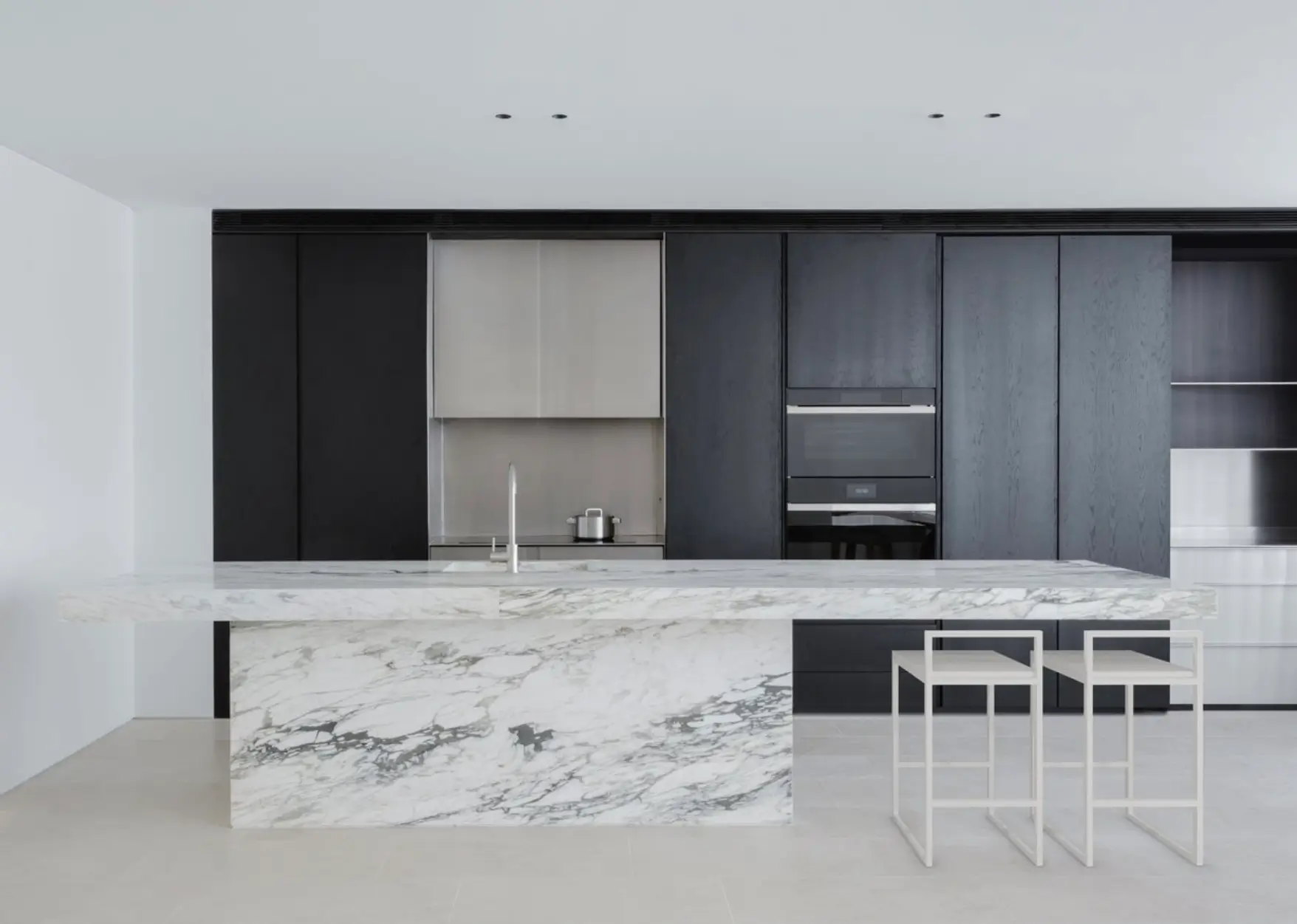 Minimalist contemporary kitchen with black floor-to-ceiling cabinetry, a sculptural white marble island, integrated appliances, and two white bar stools.