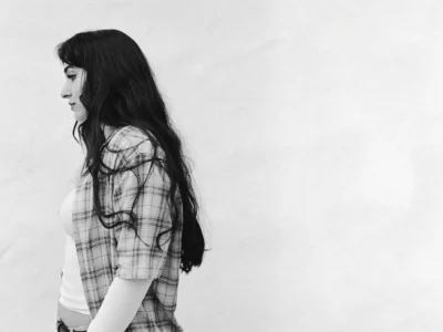 yana musician in side profile with long dark hair, wearing a plaid short-sleeve shirt over a white long-sleeve top and jeans, standing against a plain white wall with handwritten blue text reading “Come On” and “Yana” repeated across the top