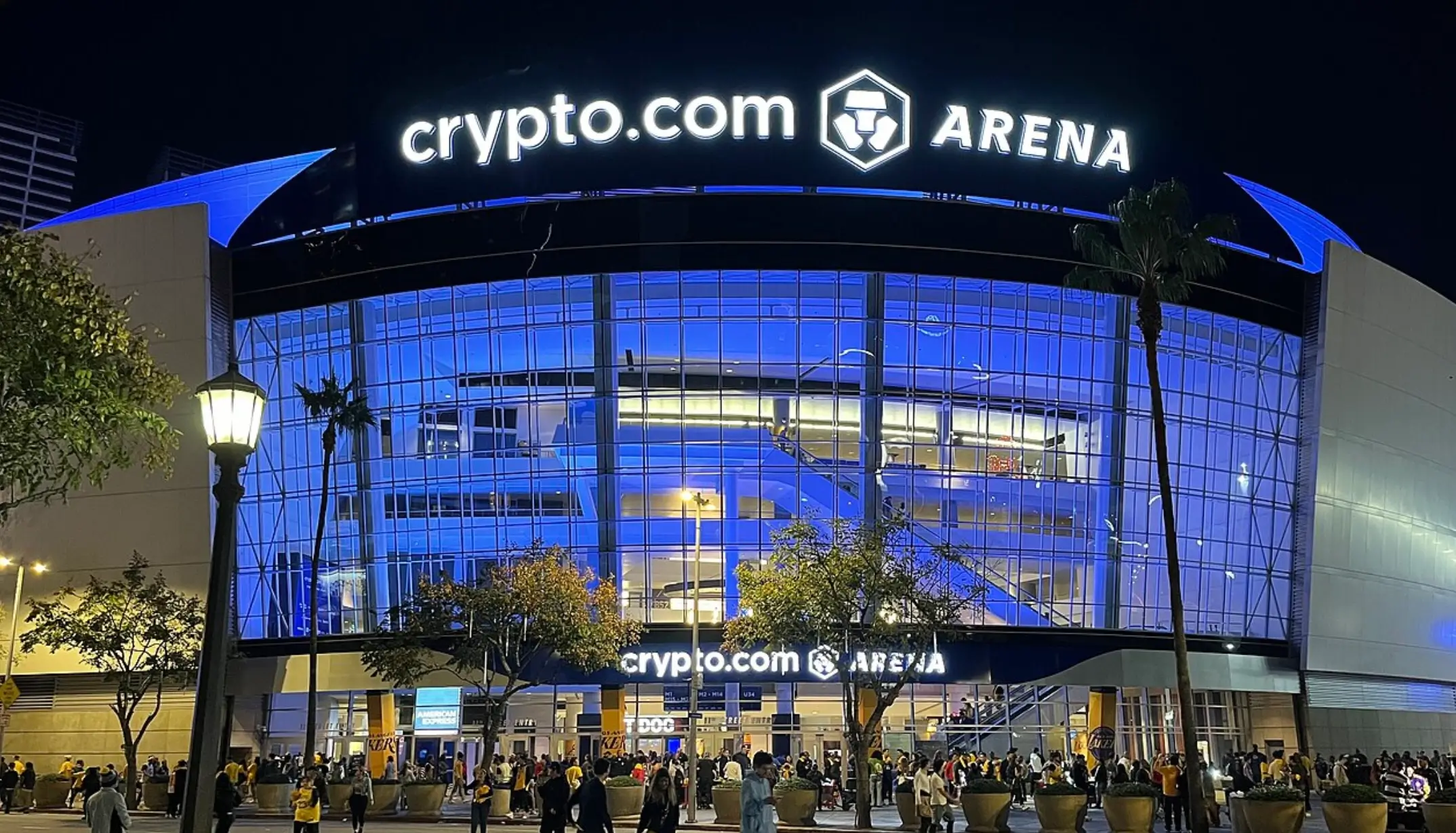 Nighttime Exterior Of Crypto.com Arena In Los Angeles, Illuminated In Blue With Its Glass Facade, Bright Arena Signage, Palm Trees And Crowds Gathering Outside The Entrance.