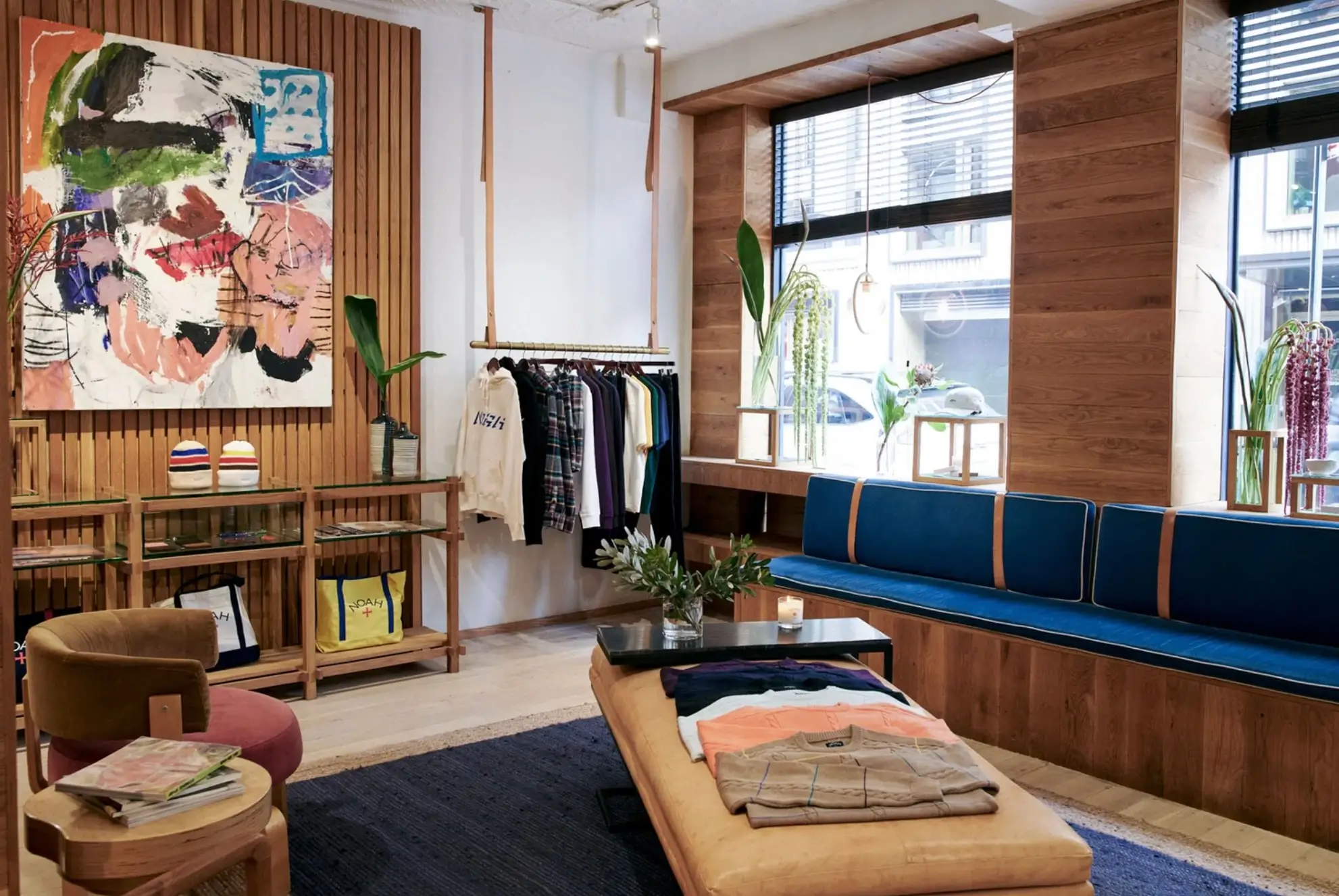 Interior Of A Noah Retail Store Featuring Warm Wood Paneling, Clothing Displays, Abstract Artwork, Blue Window Seating, Plants, And Folded Apparel.