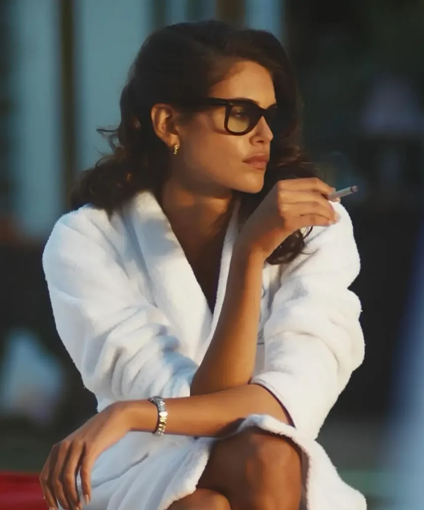 Kaia Gerber Jordan in a white robe wear oversized black sunglasses and gold jewelry, seated with one arm crossed while hold a cigarette near her face