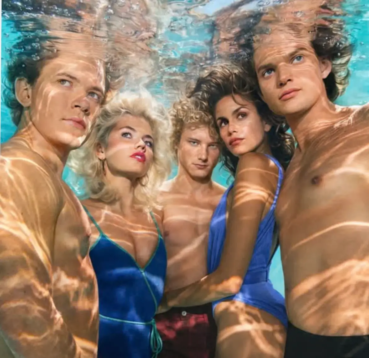 Five models posing underwater in blue swimwear, surrounded by turquoise water and rippling sunlight reflections.
