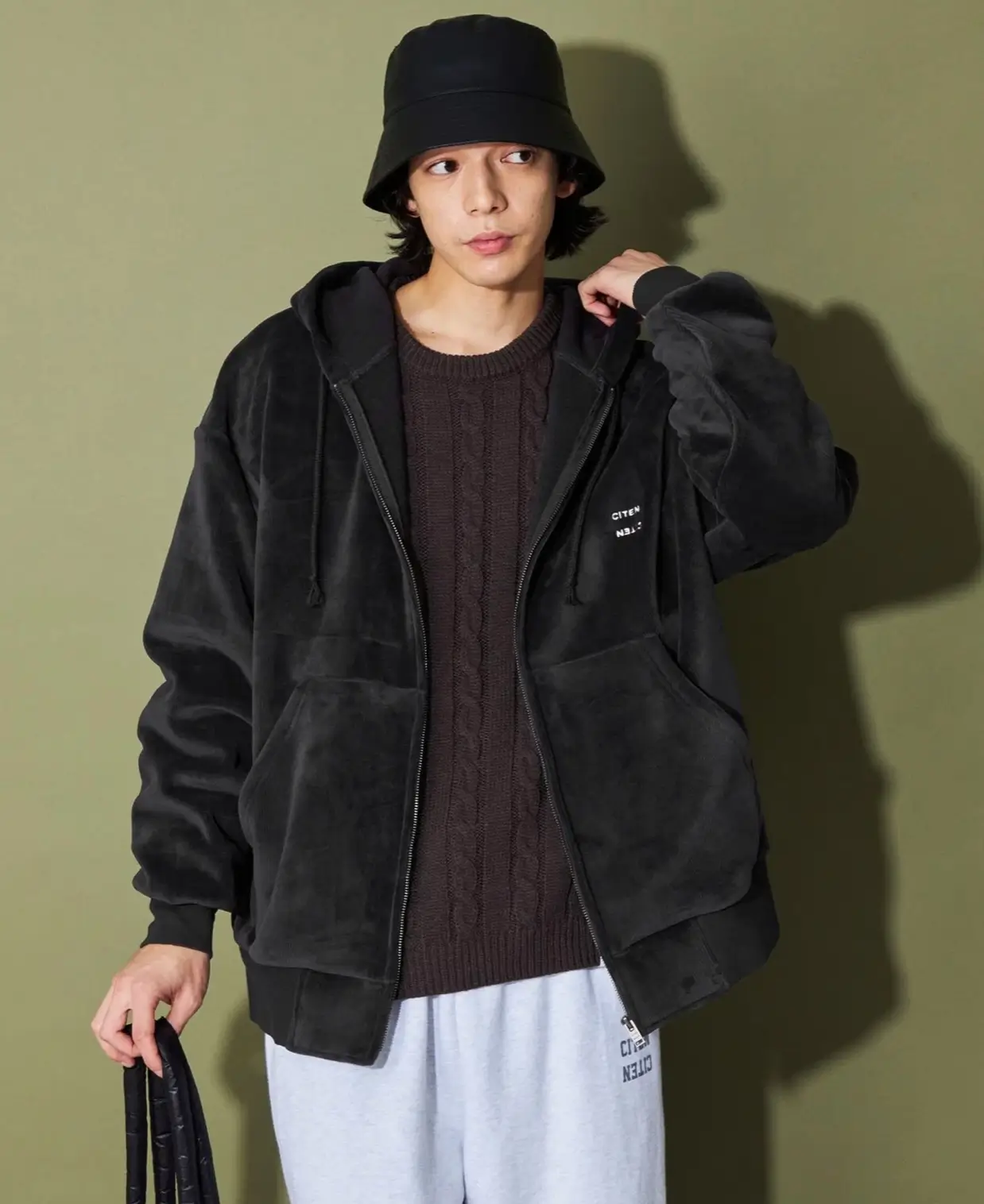 Model Wearing Black Bucket Hat And Oversized Black Zip Hoodie Over A Brown Cable-Knit Sweater