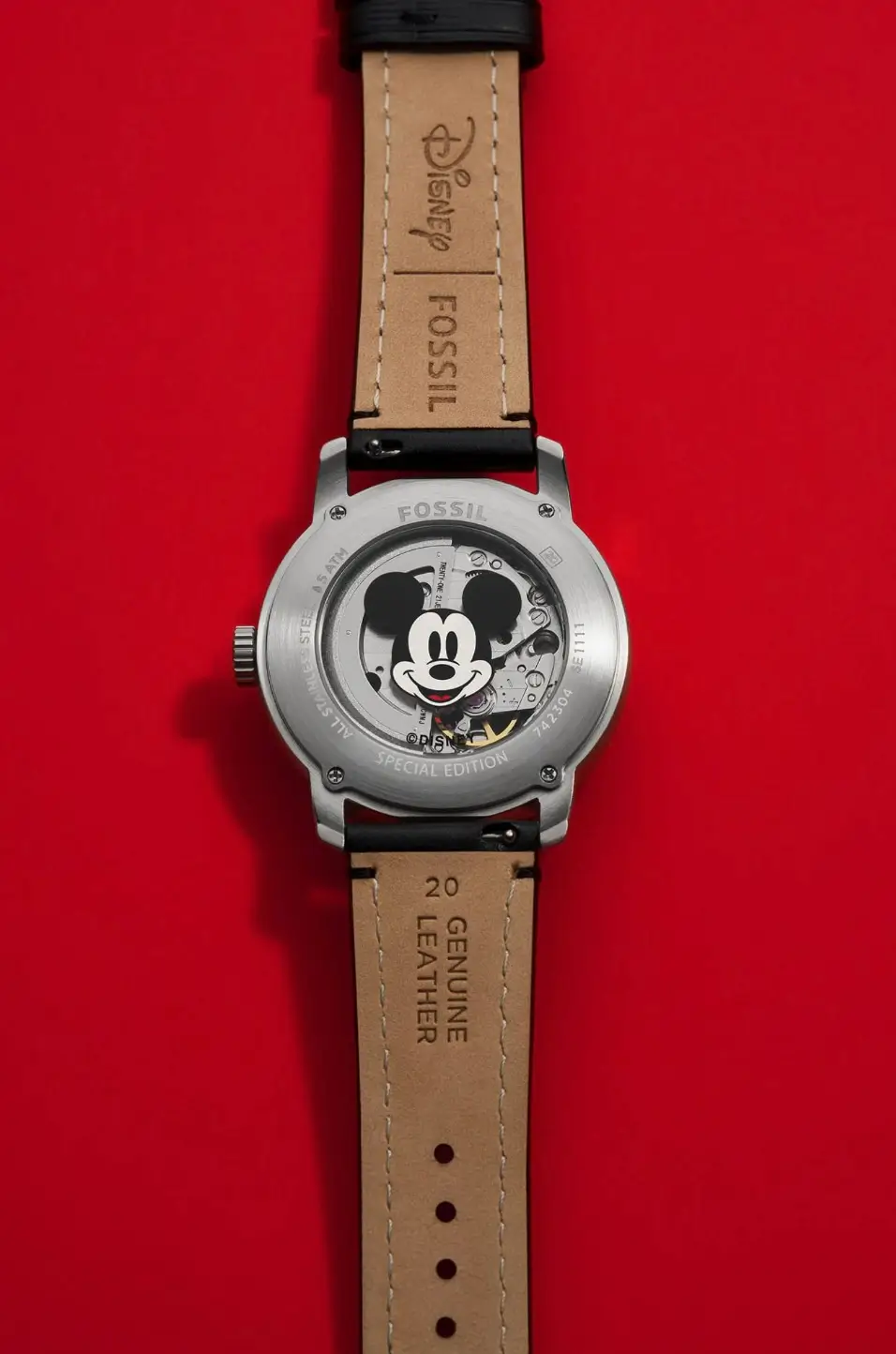 Fossil Mickey Mouse Special-Edition Watch Reveals Its Mechanical Caseback And Leather Strap Against A Red Background.