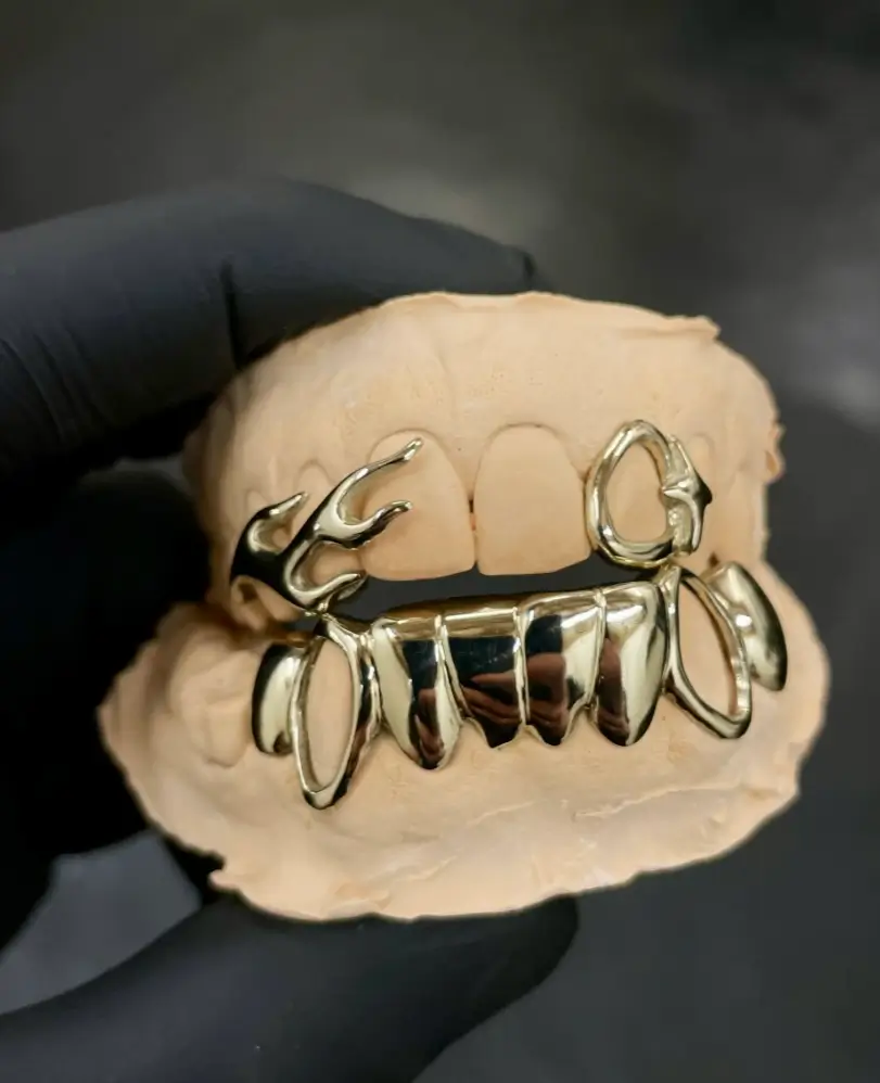Black-gloved hand holds a dental mold fitted with sculptural silver grills featuring fang-like forms and organic loops.