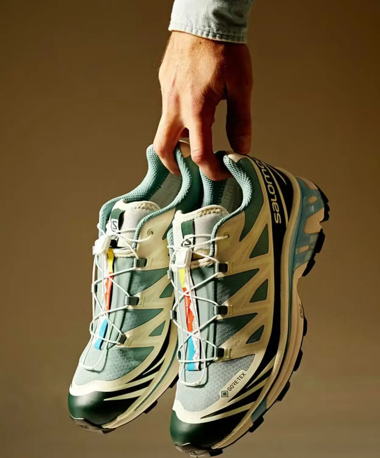 Hand Holds Pair Of Green And Cream Salomon Gore-Tex Trail Shoes With Quicklace System