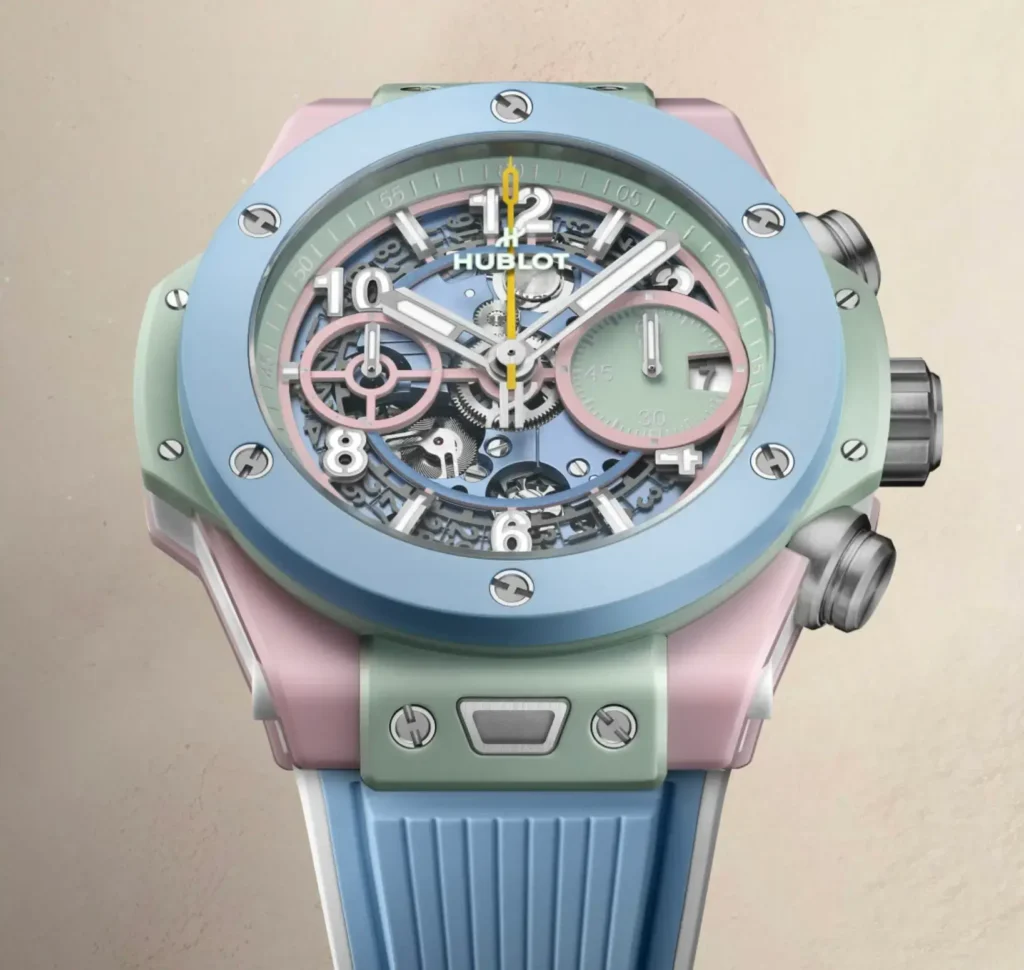 Hublot Big Bang watch appears in a close frontal view with a pastel blue ceramic bezel, mint and pink case, expos mechanical dial, and blue strap against a warm beige background