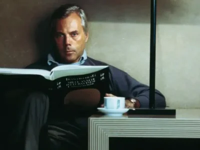 Fersan Ozpetek seated in a minimalist lounge reading a large book beside a modern table lamp, with a white coffee cup on a geometric side table and warm, subdued light
