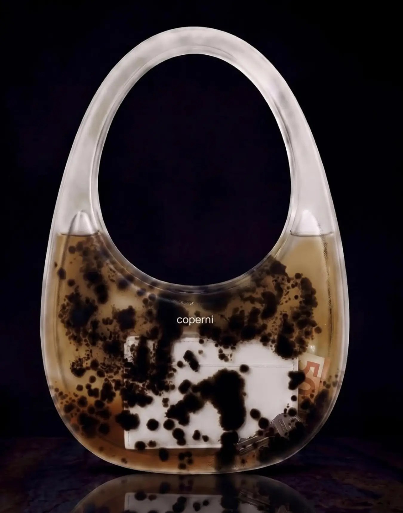 A transparent Coperni swipe bag filled with amber liquid and suspended black organic growths, revealing everyday items inside against a black background.