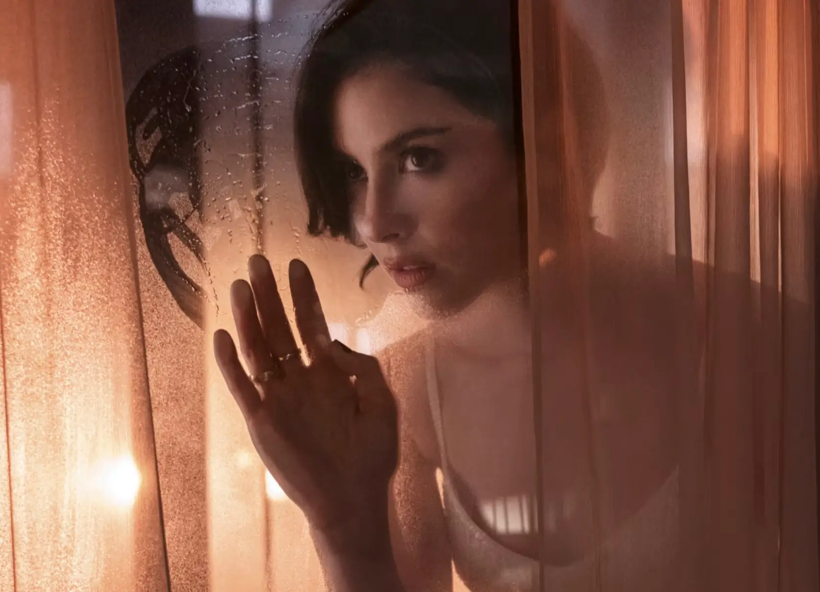 Gracie Abrams peers through sheer curtains, raising one hand in warm amber light