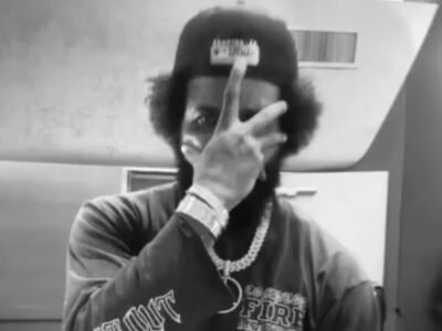The Game Wear A Backward Cap, Chain Necklace, And Watch While Hold His Hand Across His Face In A Black-And-White Portrait
