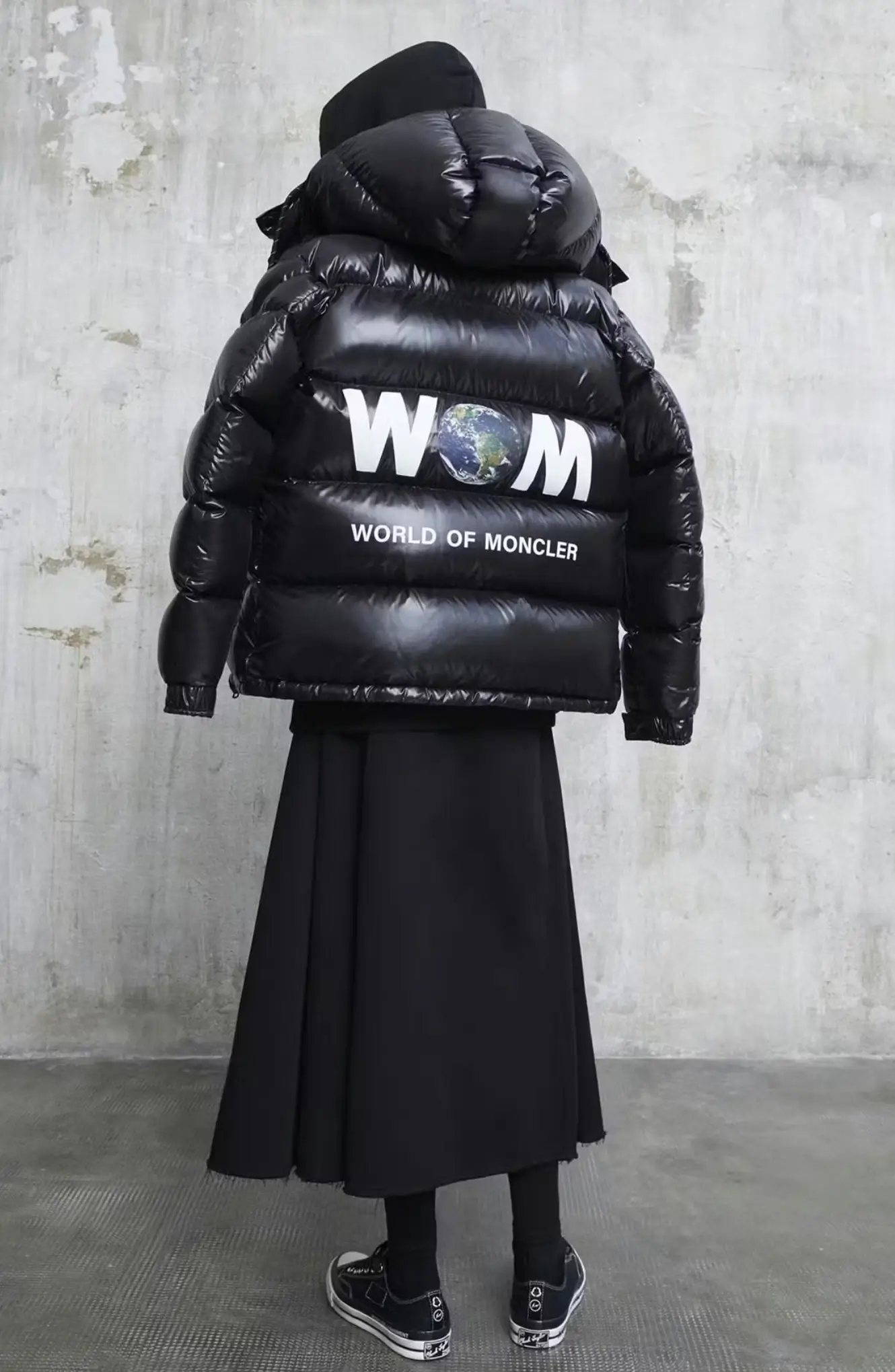 Person wearing a black puffer jacket with “WOM” graphic and long black skirt, standing against a concrete wall