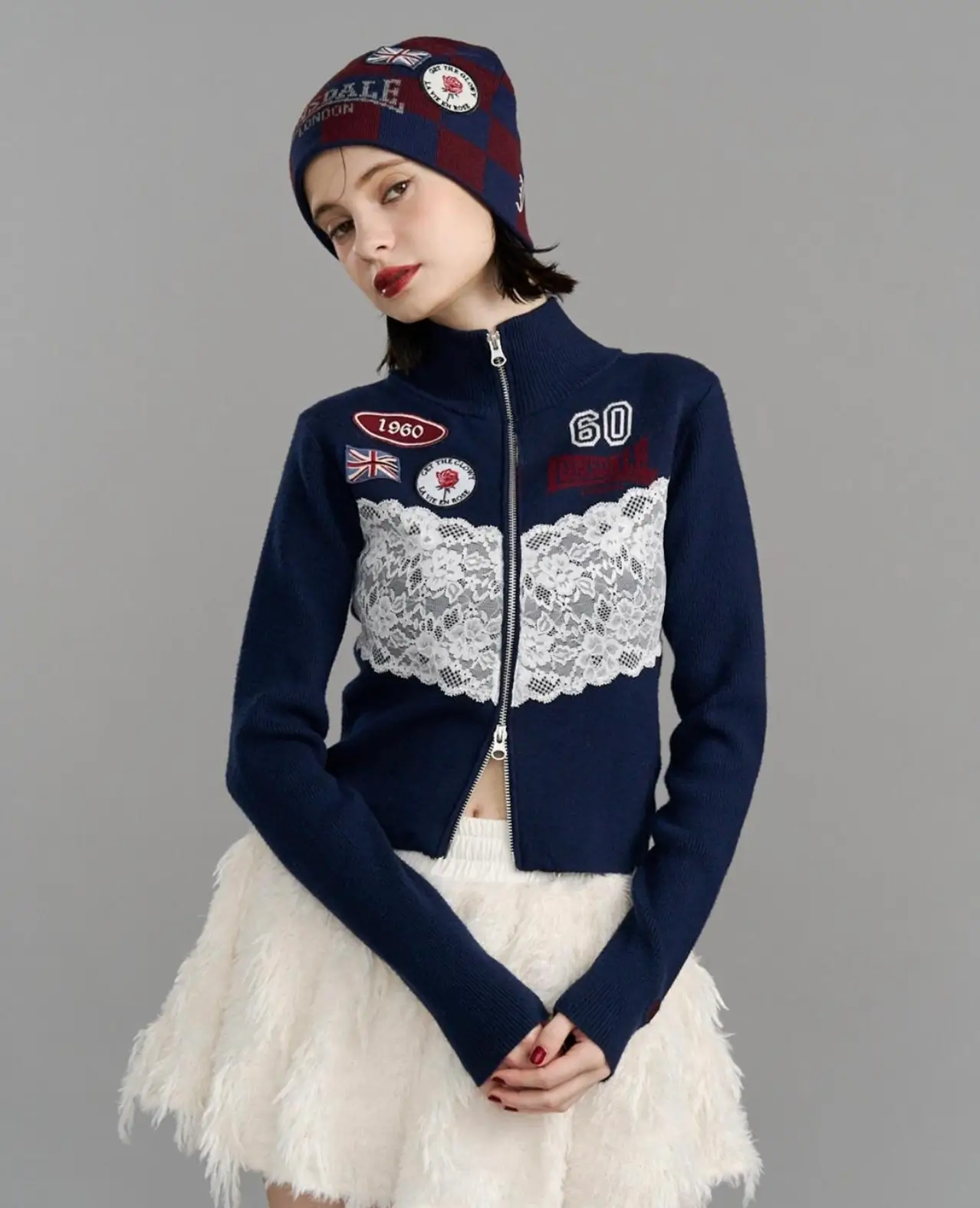 Model wearing a navy LONSDALE zip-up with lace appliqué, white furry skirt, and matching red-and-navy beanie