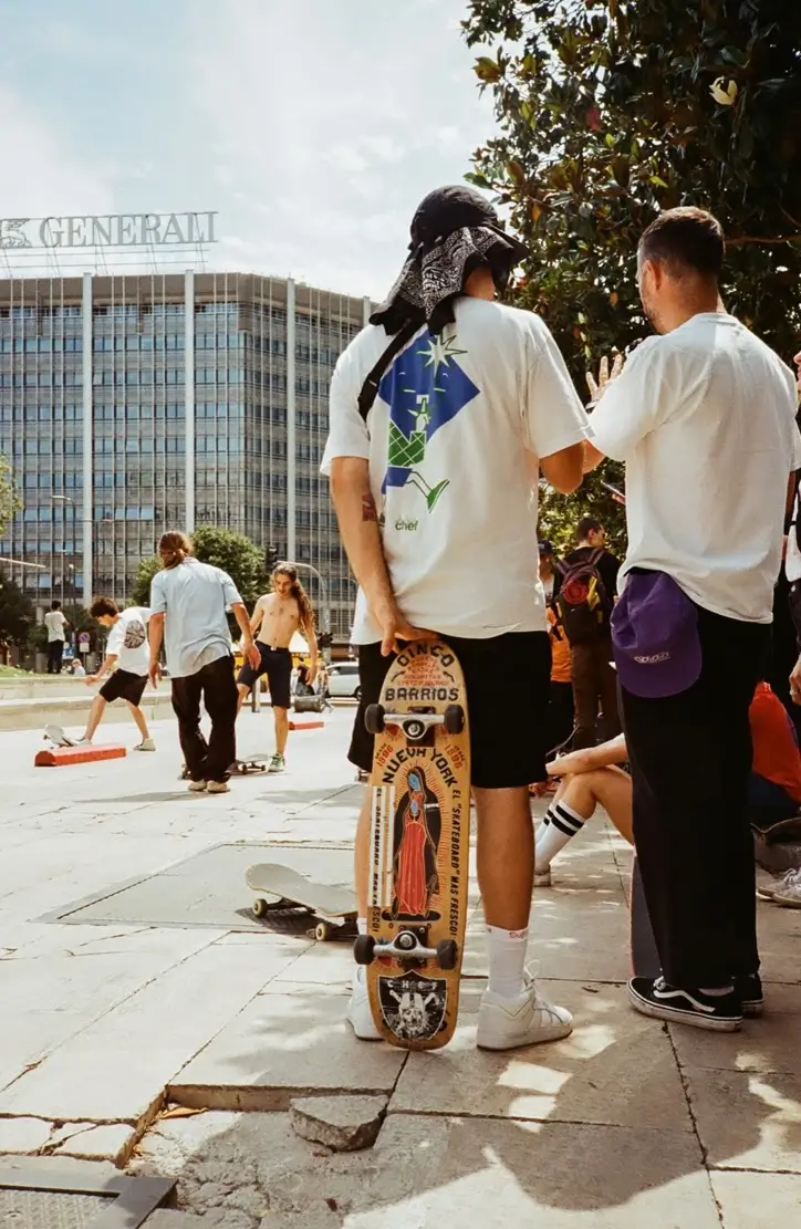 Skaters gather in a plaza; one holds a graphic skateboard and wears a bandana, with a modern building behind them