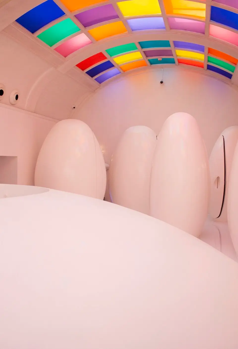 Minimalist restroom featuring oversized white egg-shaped toilet pods beneath a vaulted ceiling of colorful illuminated glass panels.