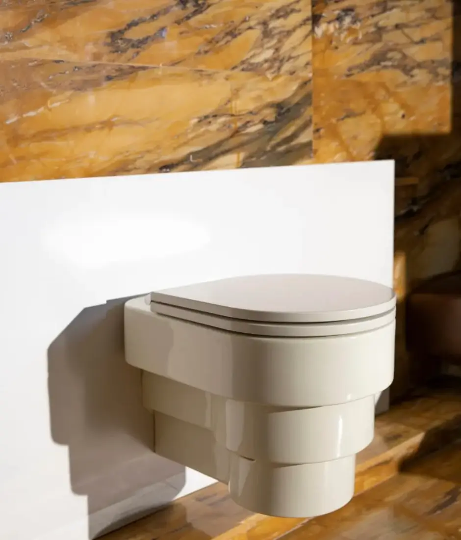 Cream-colored wall-mounted designer toilet installed against a white partition with dramatic golden marble walls and warm natural light.