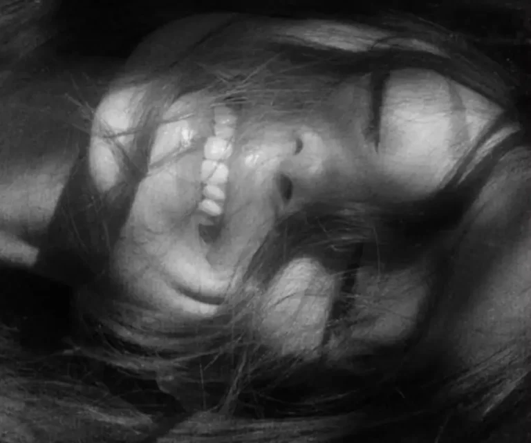 Black-and-white close-up portrait of a smiling Ariana Grande lying on their side with long hair partially covering their face, creating a soft, intimate, and dreamy composition