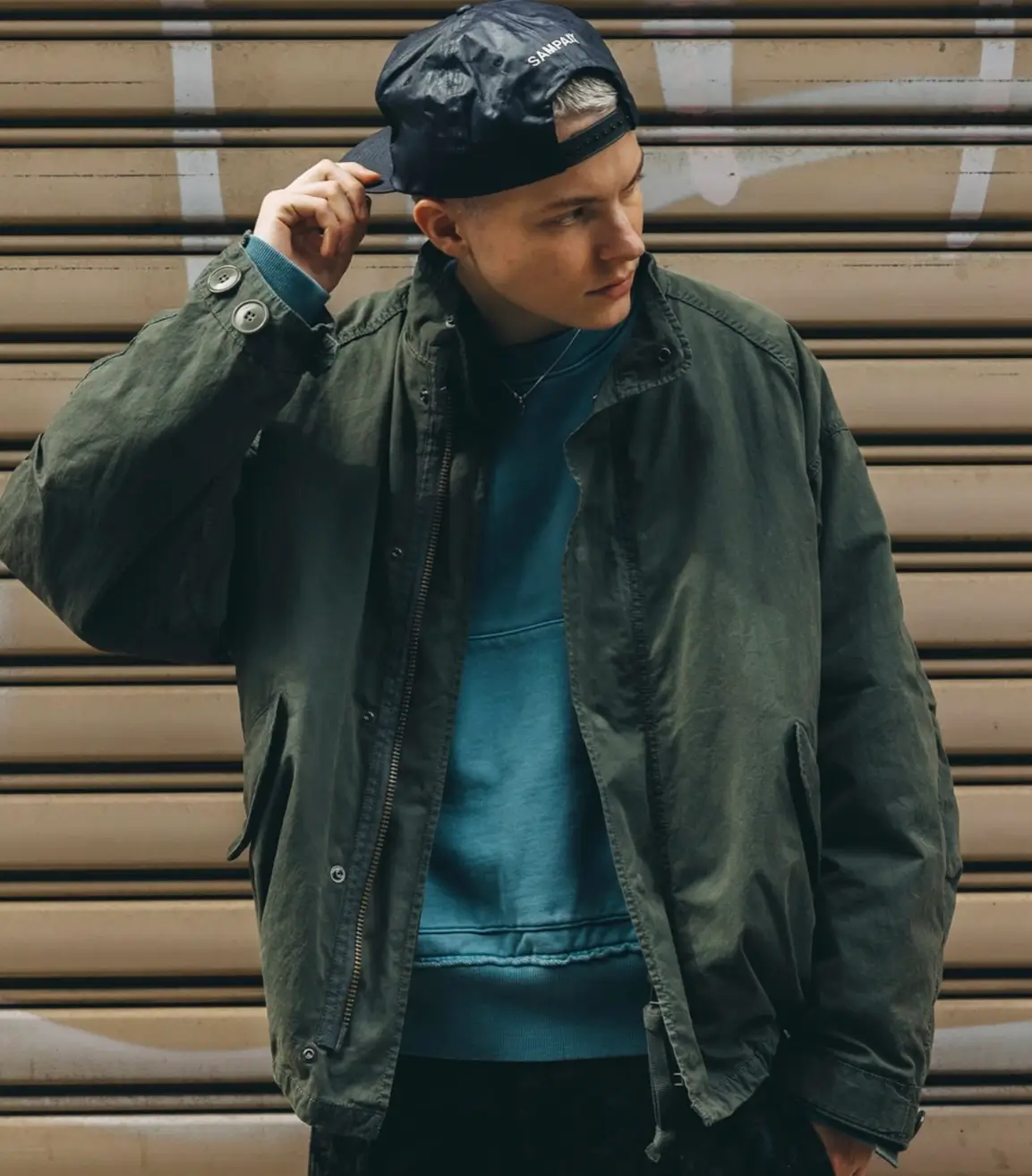 Model wearing a dark olive garment-dyed utility jacket over a teal sweatshirt with a black cap, standing in front of a metal shutter.