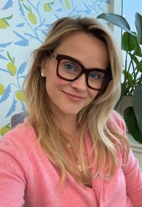 Blonde woman wearing oversized brown glasses and a pink cardigan, smiling beside indoor plants.