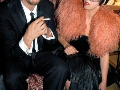 Isabella Blow right and Alexander McQueen seated in a lounge, one in a black suit and the other in a black dress with oversized blush feather shoulders and a birdcage veil
