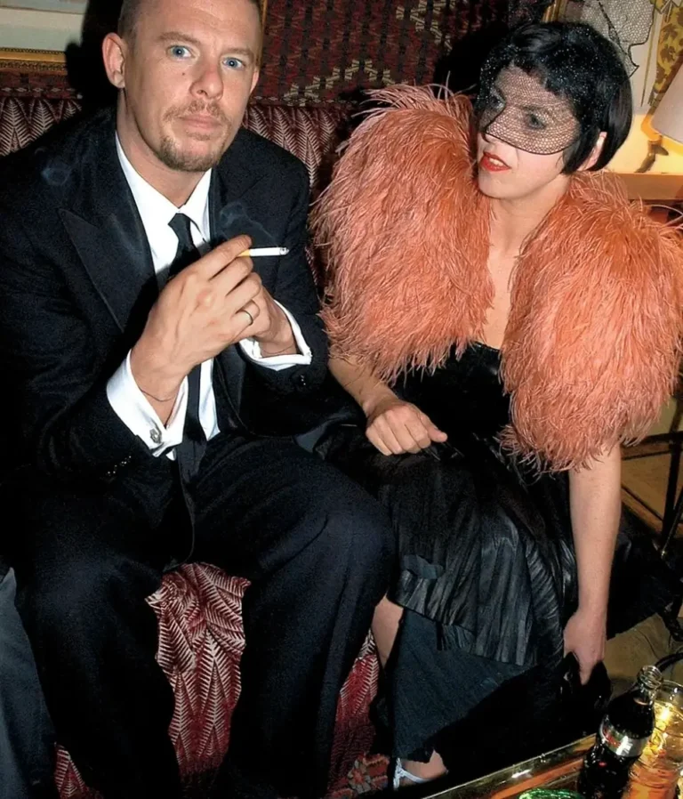 Isabella Blow right and Alexander McQueen seated in a lounge, one in a black suit and the other in a black dress with oversized blush feather shoulders and a birdcage veil