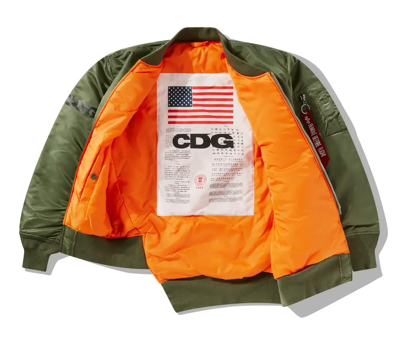 Olive green CDG MA-1 bomber jacket opened to reveal a bright orange lining with a large interior graphic featuring an American flag, CDG branding, and printed text.