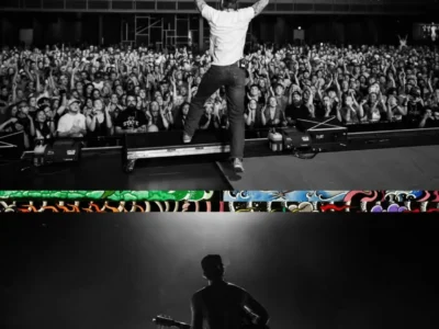 A black-and-white photograph captures a Cameron Whitcomb from behind on stage with both arms raised toward a packed concert audience. Fans fill the main floor and balcony, applauding and reaching toward the stage as venue lighting highlights the energetic live performance