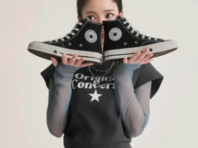 KARINA holds a pair of black Converse Chuck Taylor All Star high-top sneakers in front of their face while wearing a Converse graphic top, white skirt, and sheer sleeves against a clean studio background