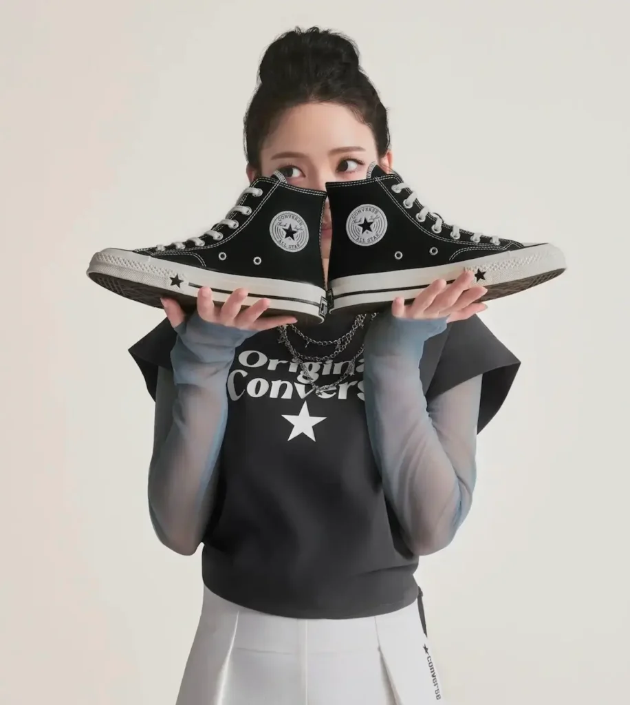 KARINA holds a pair of black Converse Chuck Taylor All Star high-top sneakers in front of their face while wearing a Converse graphic top, white skirt, and sheer sleeves against a clean studio background
