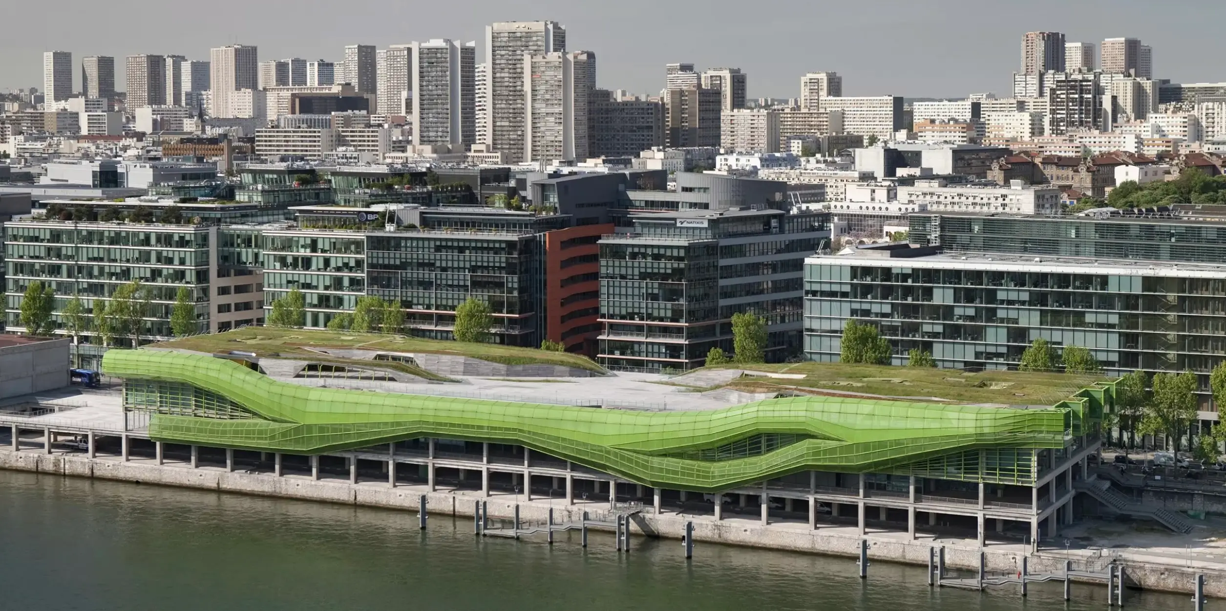 Panoramic view of a green wave-shaped riverside building surrounded by modern office blocks and a dense city skyline.