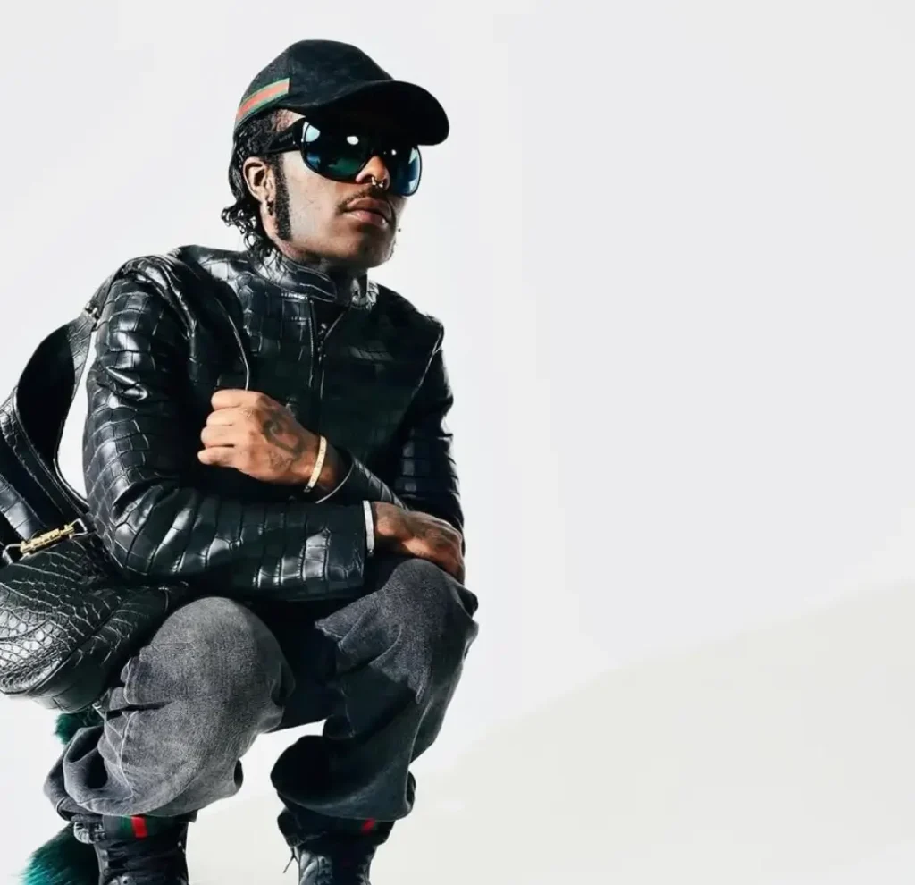 Lil Uzi Vert crouching in a black textured leather jacket, dark jeans, oversized sunglasses, and a black cap, carrying a matching black leather bag against a white background